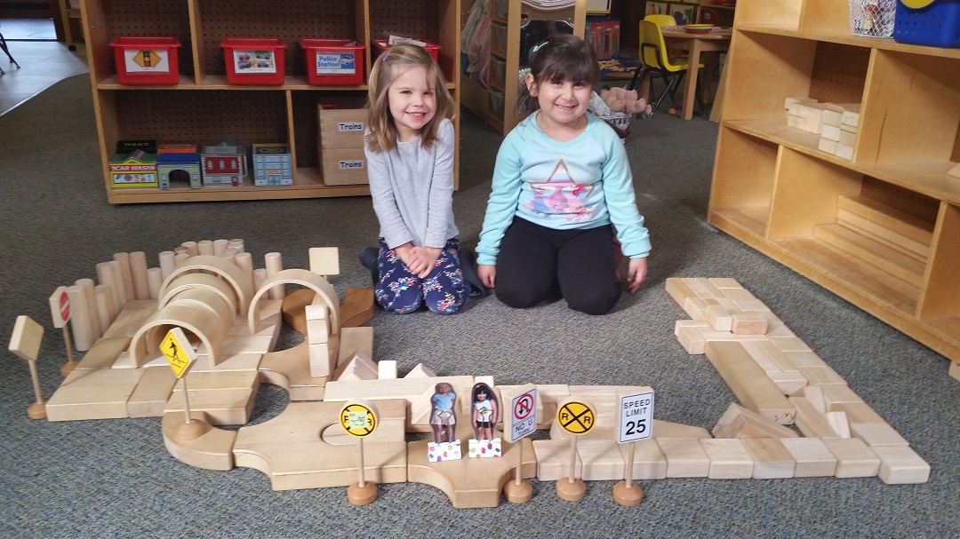 Two young girls smile beside a wooden block road setup with traffic signs in a classroom.